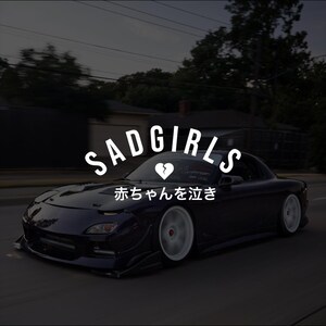 Puede incluir: Un coche deportivo azul oscuro con ruedas blancas está en movimiento en una carretera. El texto "SADGIRLS" está encima del coche, con un símbolo de corazón roto. Debajo hay texto japonés.