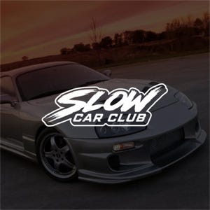 Op de afbeelding: Een zilverkleurige sportwagen met een luchtinlaat in de voorbumper en vijfspaaks wielen. Op de motorkap staat een wit grafisch logo in een penseelstijl lettertype met de tekst SLOW CAR CLUB in dikke hoofdletters met een zwarte omlijning.