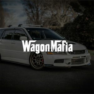 Peut inclure: Une voiture break blanche avec le texte "Wagon Mafia" en blanc. La voiture a un capot noir, des vitres teintées et une galerie de toit. La voiture est garée sur une route asphaltée sombre.
