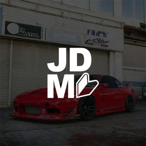May include: A red sports car with black wheels and a spoiler is parked in front of a garage. The white text "JDM" is overlaid on the image. The car has a sleek design and appears to be modified.
