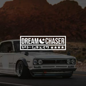 Peut inclure: Autocollant rectangulaire blanc avec le texte "DREAM CHASER" au-dessus de caractères japonais et d'étoiles. L'autocollant est affiché sur le devant d'une voiture vintage blanche avec des accents noirs, garée sur une route avec une montagne en arrière-plan.