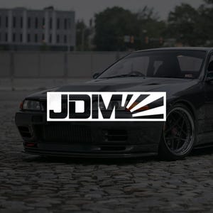 Peut inclure: Une voiture de sport noire avec le texte "JDM" dans un rectangle blanc. La voiture est garée sur une rue pavée. Le logo présente un motif de soleil levant.