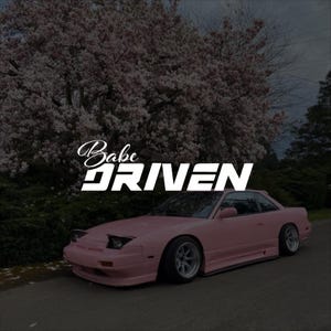 Puede incluir: Un coche deportivo rosa con el texto "Babe Driven" en blanco. El coche está estacionado en una carretera pavimentada con un fondo de un árbol en flor. El coche tiene un diseño elegante con ruedas personalizadas.