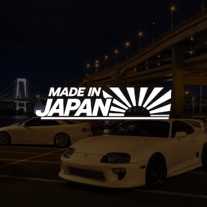 Peut inclure: Une voiture de sport blanche garée de nuit avec une superposition graphique présentant le texte MADE IN JAPAN et un motif de soleil stylisé. La scène comprend une deuxième voiture blanche en arrière-plan et un pont éclairé par des réverbères.