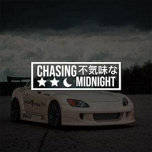 May include: A cream-coloured sports car with a rear spoiler, parked on tarmac. A white rectangular sticker with the words "CHASING 不気味な MIDNIGHT" and star and crescent moon graphics is above the car.