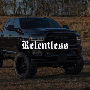 Peut inclure: Un pick-up noir avec de gros pneus est garé sur un champ herbeux. Le mot "Relentless" est affiché en blanc, dans un texte stylisé sur le devant du camion. Le camion a une calandre et des phares noirs.