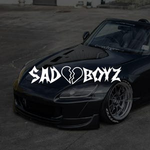 May include: Black convertible car with a white "SADBOYZ" decal featuring a broken heart graphic on the bonnet. The car has silver rims and low-profile tyres. The image is taken outdoors on a paved surface.