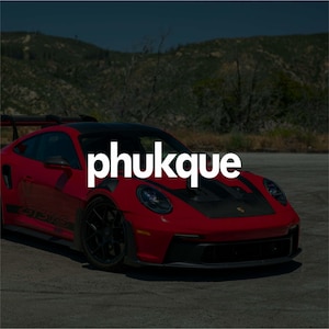 May include: A red sports car with black accents and the text "phukque" in white. The car is parked on a paved road with a mountain backdrop. The car has black wheels and a spoiler.