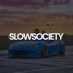 May include: A blue sports car with a custom body kit and a rear wing is parked on a paved surface. The text "SLOWSOCIETY" is displayed in white across the top of the image. The sky is a mix of orange and purple.