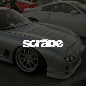 Puede incluir: Un coche deportivo plateado con la palabra "scrape" en blanco en el capó. El coche tiene un perfil bajo, llantas personalizadas y está aparcado cerca de otros vehículos. La imagen está tomada en un ligero ángulo, mostrando el diseño elegante del coche.