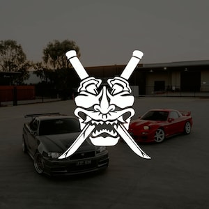 Könnte beinhalten: Weißes Grafikdesign einer japanischen Dämonenmaske mit gekreuzten Schwertern. Links ist ein dunkelgraues Sportwagen geparkt, rechts ein roter Sportwagen. Der Hintergrund ist ein Parkplatz mit einem Gebäude und Bäumen.