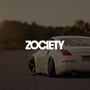 Op de afbeelding: Een witte sportwagen met zwarte velgen en het woord "Zociety" in witte tekst. De auto staat op een weg met een vage achtergrond van bomen en een schemerige lucht. De achterlichten van de auto zijn rood.