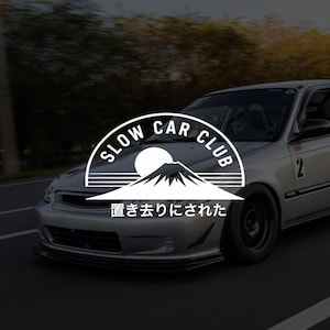 Peut inclure: Une berline argentée roulant sur une route avec un logo circulaire blanc superposé au centre. Le logo présente le texte SLOW CAR CLUB, un soleil, une silhouette de montagne et le texte japonais 置き去りにされた. La voiture a une lèvre avant noire.