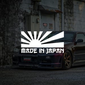 May include: A dark-coloured car with a white graphic of a rising sun and the text "MADE IN JAPAN". The car is parked in front of a weathered brick wall.
