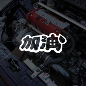 May include: A car engine bay featuring a red engine cover and various mechanical parts. A white sticker with Japanese characters and a drop graphic is overlaid on the image. The engine bay is dark, highlighting the engine's details.