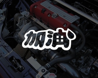 May include: An engine bay with a red engine cover and various mechanical components. A white sticker with Japanese characters and a drop graphic is overlaid on the image. The engine bay is dark, with a focus on the engine's details.