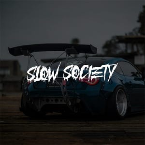 Peut inclure: Une voiture de sport bleu sarcelle avec un grand aileron noir et un échappement personnalisé. Le texte "SLOW SOCIETY" est affiché en blanc, avec une police dégoulinante sur l'arrière de la voiture. La voiture est garée sur une surface en bois.