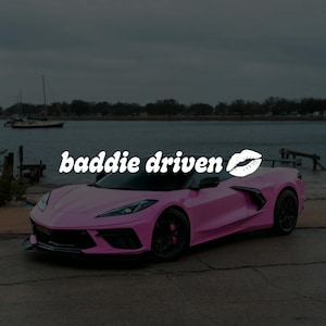 Op de afbeelding: Een roze sportwagen met zwarte wielen en bekleding staat geparkeerd bij een water. De auto heeft de tekst "baddie driven" in het wit over de voorruit, met een witte lip-afbeelding.