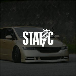 May include: A gold car with black wheels and a lowered suspension. The word "STATIC" is displayed in large white letters across the windscreen, with a graphic of a shock absorber integrated into the letter "C".