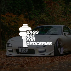 May include: A white sports car with the text "BAGS ARE FOR GROCERIES" in white. The car is parked on a surface covered in fallen leaves. The car has a license plate.