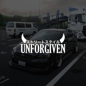 May include: A black car with the word "UNFORGIVEN" in white block letters. Above the word is Japanese text and a white horn-like graphic. The car is parked in a car park with a white van and a lorry in the background.