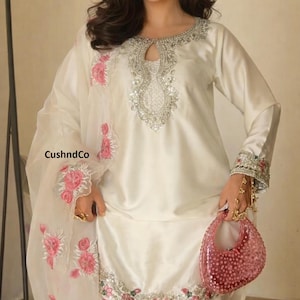 May include: Ivory-colored satin ensemble featuring a long-sleeved tunic with intricate silver and crystal embellishments around the neckline and cuffs. The outfit includes matching pants and a sheer, floral-embroidered shawl. A pink, beaded handbag completes the look. The text "CushndCo" is visible.