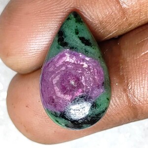 May include: A teardrop-shaped gemstone with a striking pink floral centre, framed by green and black. The polished stone is held in a hand, highlighting its natural beauty and unique colour combination.