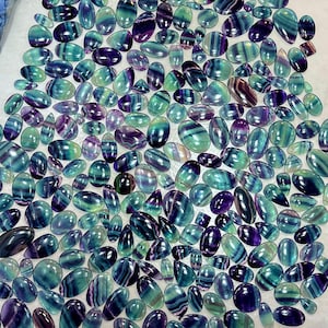 May include: A collection of polished fluorite gemstones in various shapes and sizes. The stones display a mix of colors, including shades of green, blue, and purple, with striped patterns. The gemstones are oval, teardrop, and other shapes.