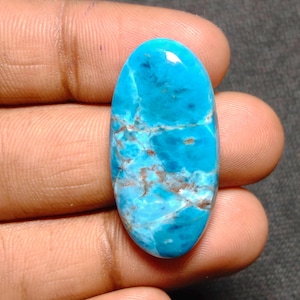 Puede incluir: Una gema ovalada de un vibrante color turquesa, con intrincados patrones de vetas marrones y blancas. La superficie pulida refleja la luz, resaltando la belleza natural de la piedra. Esta gema es un ejemplo llamativo de artesanía en piedra natural.