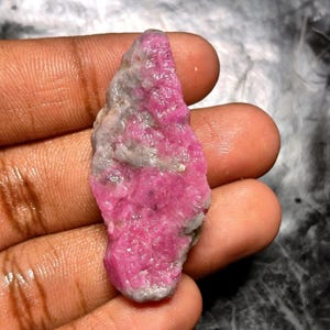 May include: A rough, uncut ruby gemstone with a vibrant pink hue, held in a hand. The ruby displays a mix of pink and gray tones, showcasing its natural, raw texture. The gemstone is irregularly shaped, highlighting its organic origin.