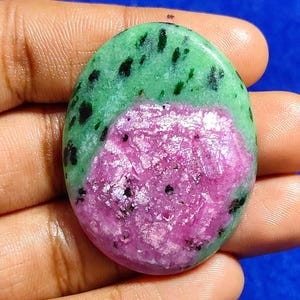 Puede incluir: Gema ovalada con una vibrante combinación de colores. La mitad superior es de un verde intenso con motas negras, que se transforma en un llamativo tono rosa-rojizo en la parte inferior. La superficie pulida refleja la luz, resaltando la belleza natural de la piedra.