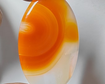 May include: Oval-shaped gemstone with a vibrant orange and white pattern. The polished stone has a glossy finish, with the orange color swirling in a circular design. The bottom half of the stone is a creamy white color.