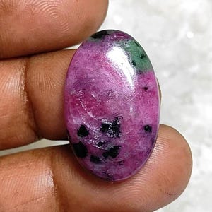Puede incluir: Gema de forma ovalada con superficie pulida. La piedra presenta un vibrante tono rosa-rojizo, acentuado con parches negros y verdes. La gema se sostiene entre dos dedos.
