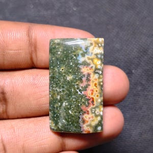 May include: A rectangular polished ocean jasper gemstone cabochon featuring a deep forest green base with intricate white star-like patterns and a distinct side band of vibrant orange, yellow, and white circular orbicular inclusions.