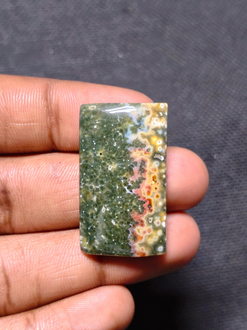 May include: A rectangular polished ocean jasper gemstone cabochon featuring a deep forest green base with intricate white star-like patterns and a distinct side band of vibrant orange, yellow, and white circular orbicular inclusions.