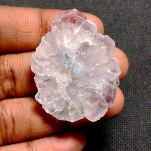 May include: A raw, uncut amethyst crystal cluster. The crystal features a pale lavender hue with a central pattern of white and green. The edges of the crystal are jagged and translucent, with a few darker purple tips.