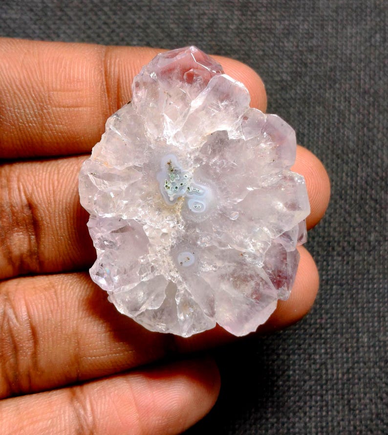 May include: A raw, uncut amethyst crystal cluster. The crystal features a pale lavender hue with a central pattern of white and green. The edges of the crystal are jagged and translucent, with a few darker purple tips.