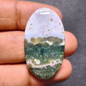 May include: An oval-shaped polished gemstone featuring a smooth surface with a distinct color gradient. The top section is a pale lavender with small circular inclusions, transitioning into a deep forest green base with white and pink spots.