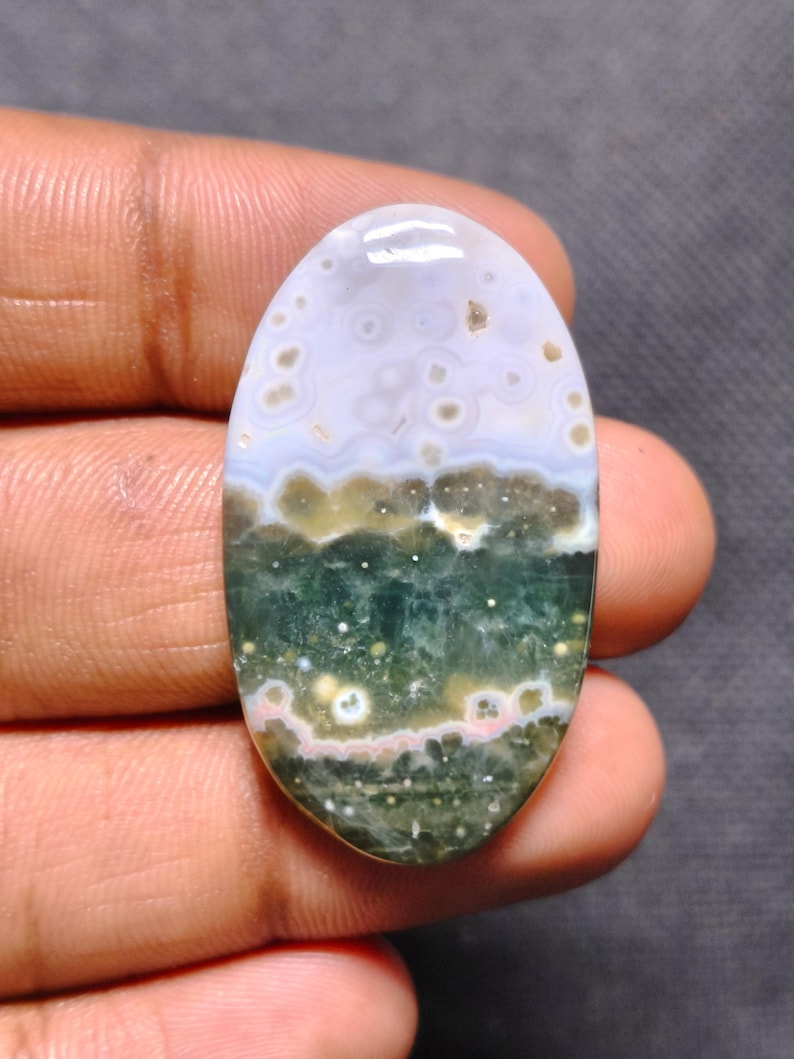May include: An oval-shaped polished gemstone featuring a smooth surface with a distinct color gradient. The top section is a pale lavender with small circular inclusions, transitioning into a deep forest green base with white and pink spots.