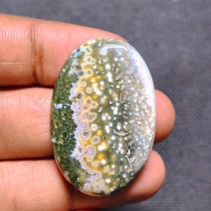 May include: An oval-shaped polished gemstone featuring a complex pattern of moss green, mustard yellow, and white circular inclusions. The stone has a smooth, glossy surface with a distinct color gradient transitioning from deep green to light.