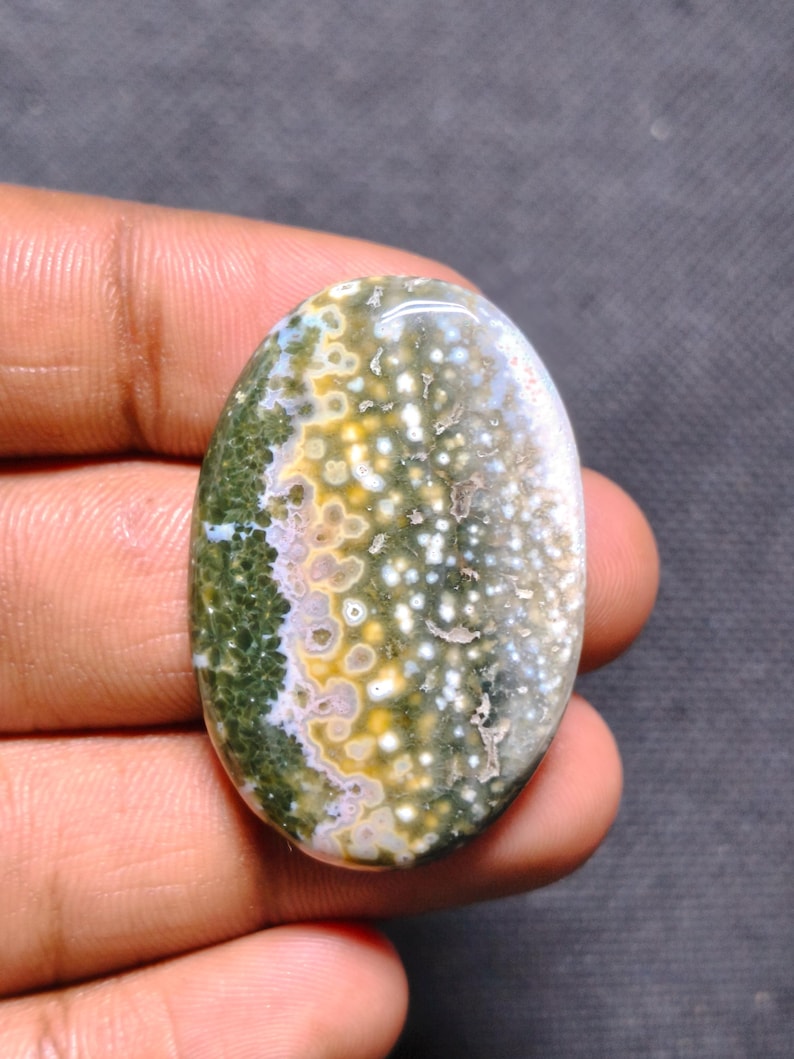 May include: An oval-shaped polished gemstone featuring a complex pattern of moss green, mustard yellow, and white circular inclusions. The stone has a smooth, glossy surface with a distinct color gradient transitioning from deep green to light.