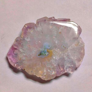 May include: A polished, irregular-shaped amethyst geode slice. The crystal features a mix of lavender, white, and clear quartz with a central pattern of blue and yellow hues. The geode has a natural, organic appearance.