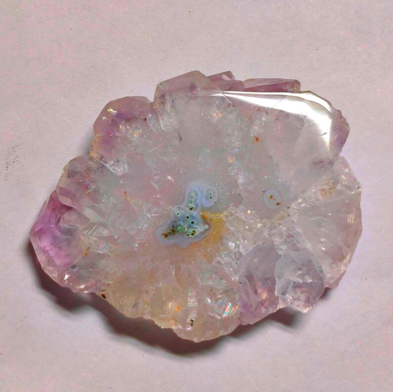 May include: A polished, irregular-shaped amethyst geode slice. The crystal features a mix of lavender, white, and clear quartz with a central pattern of blue and yellow hues. The geode has a natural, organic appearance.