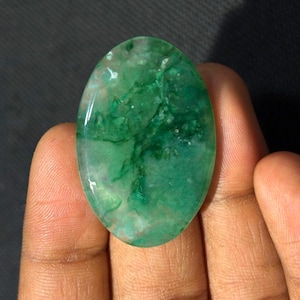 May include: An oval-shaped, polished gemstone with a translucent appearance, featuring a vibrant green color with internal white and darker green veining patterns throughout the smooth, rounded surface of the stone.