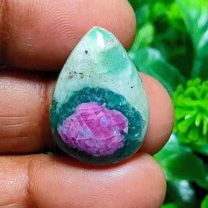 May include: A teardrop-shaped gemstone with a vibrant color palette. The gem features a central, textured magenta core, encircled by a deep teal band, and encased in a pale green exterior. The stone is held between fingers.
