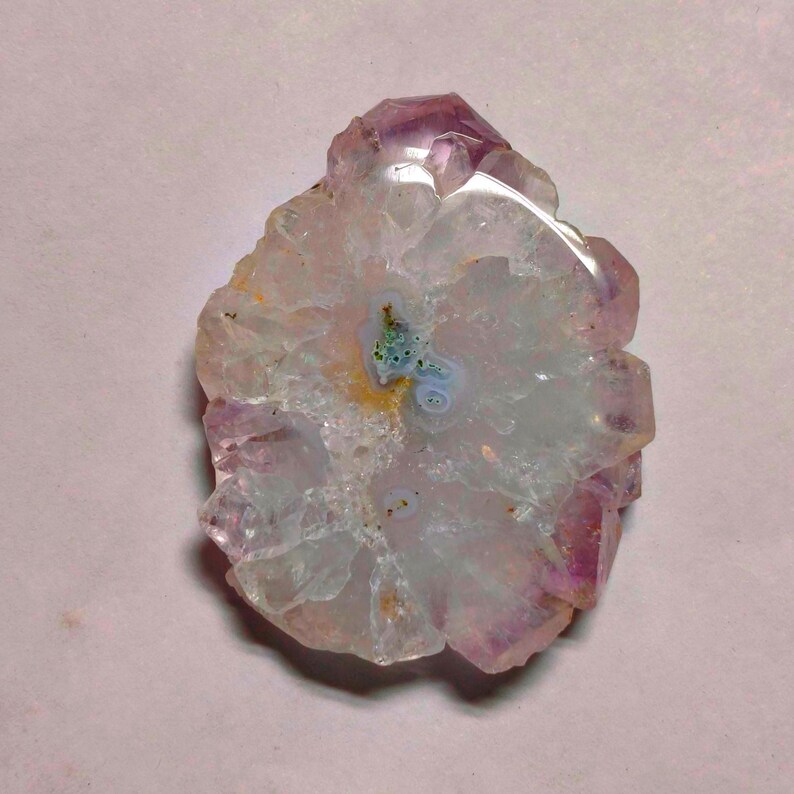 May include: A natural amethyst geode slice with a crystalline structure. The geode features a central area of pale blue and green hues, surrounded by a ring of translucent white and a border of deep purple crystals. The overall shape is irregular.