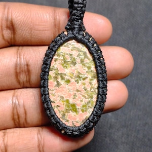 Puede incluir: Colgante ovalado con una piedra moteada en tonos rosa y verde, engastada en un diseño de macramé negro. El colgante tiene un lazo en la parte superior para colgarlo. El patrón de la piedra es único y el macramé añade un toque artesanal.