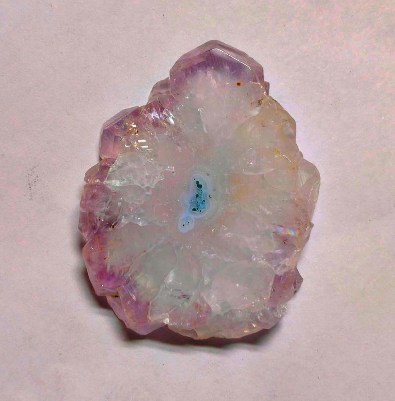May include: A polished amethyst geode slice with a central turquoise-blue core. The outer edges display a deep purple hue, transitioning to a translucent white and pink interior. The crystal formation has a natural, irregular shape.