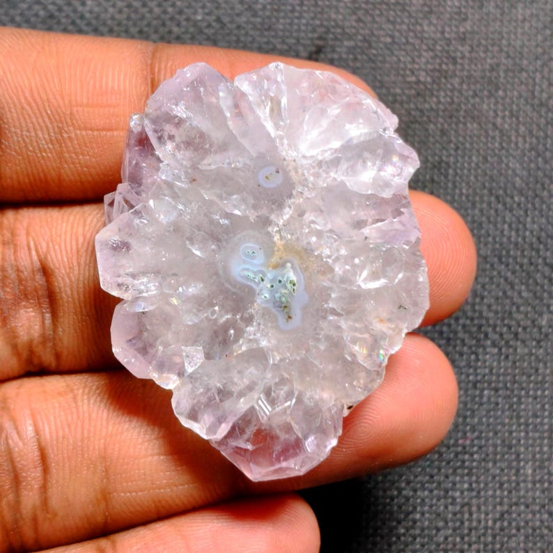 May include: A light purple and clear crystal cluster with a circular pattern in the center. The crystal has a rough, textured surface and is held in a hand. The center of the crystal has a light blue and green pattern.