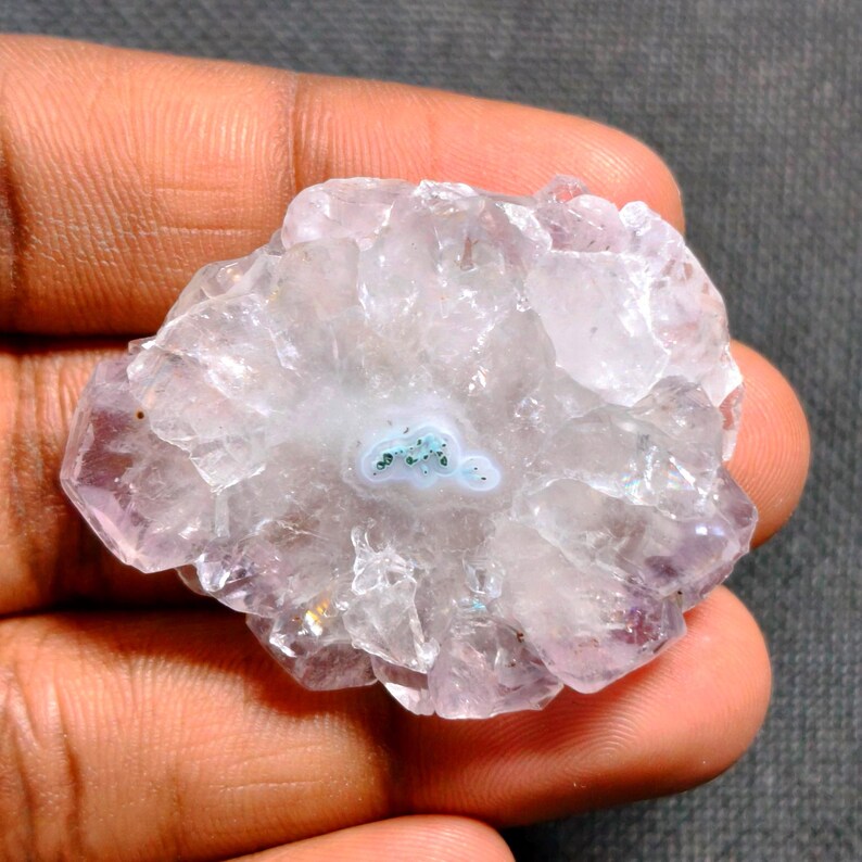 May include: A close-up of a light purple and white geode crystal. The crystal has a circular shape with a light blue center and a crystalline structure. The geode is held in a person's hand.